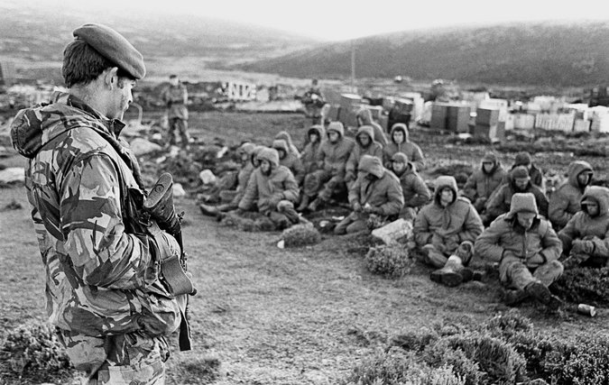 THE FALKLANDS WAR 1982: Argentinian soldiers captured at Goose Green are guarded by a British Royal Marine as they await transit out of the area.